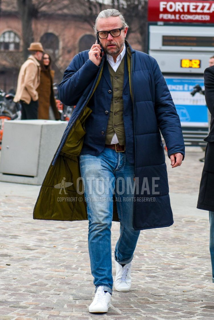 Men's coordinate and outfit with plain glasses, plain navy down jacket, plain navy tailored jacket, plain olive green gilet, plain white shirt, plain blue denim/jeans, and white low-cut sneakers.