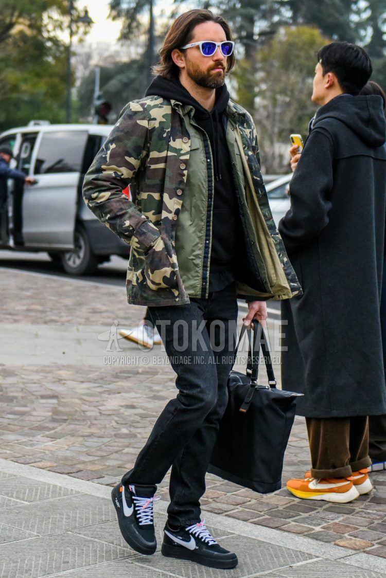 Men's coordinate and outfit with solid color sunglasses, multi-colored camouflage shirt jacket, olive green solid color coach jacket, solid color black hoodie, solid color black denim/jeans, Nike black low-cut sneakers, solid color black tote bag.