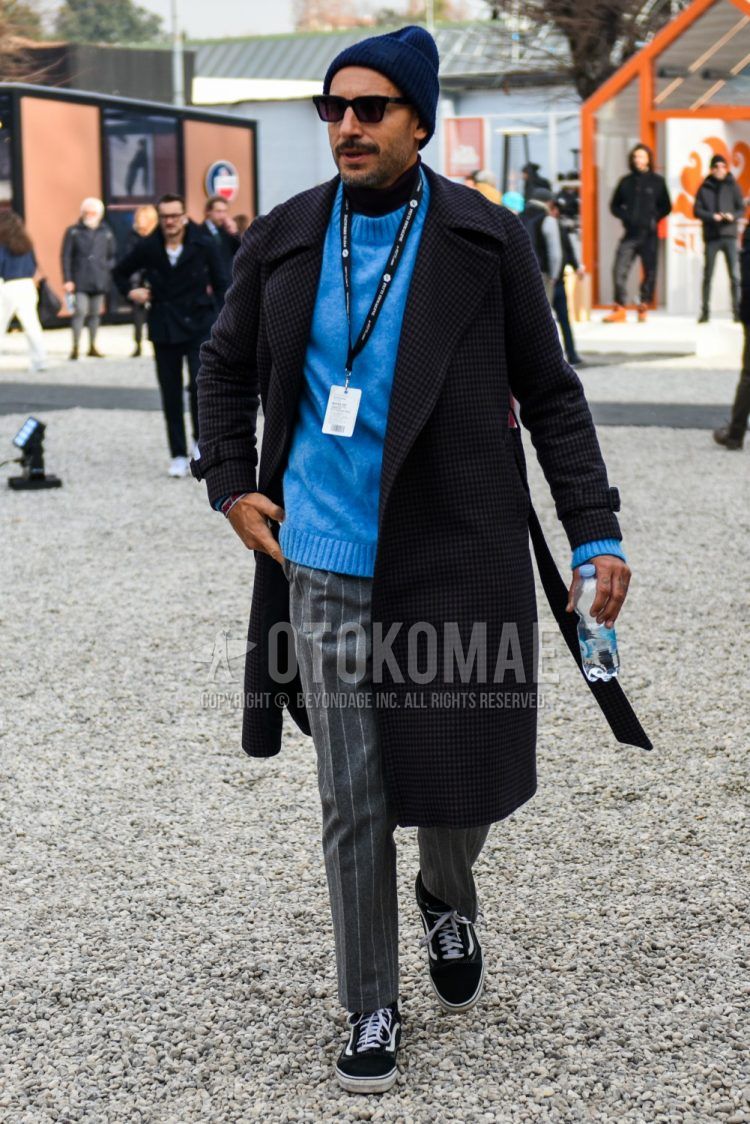 Men's coordinate and outfit with plain navy knit cap, plain sunglasses, black checked outerwear, black checked belted coat, plain blue sweater, gray striped slacks, and black low-cut sneakers by Vans.