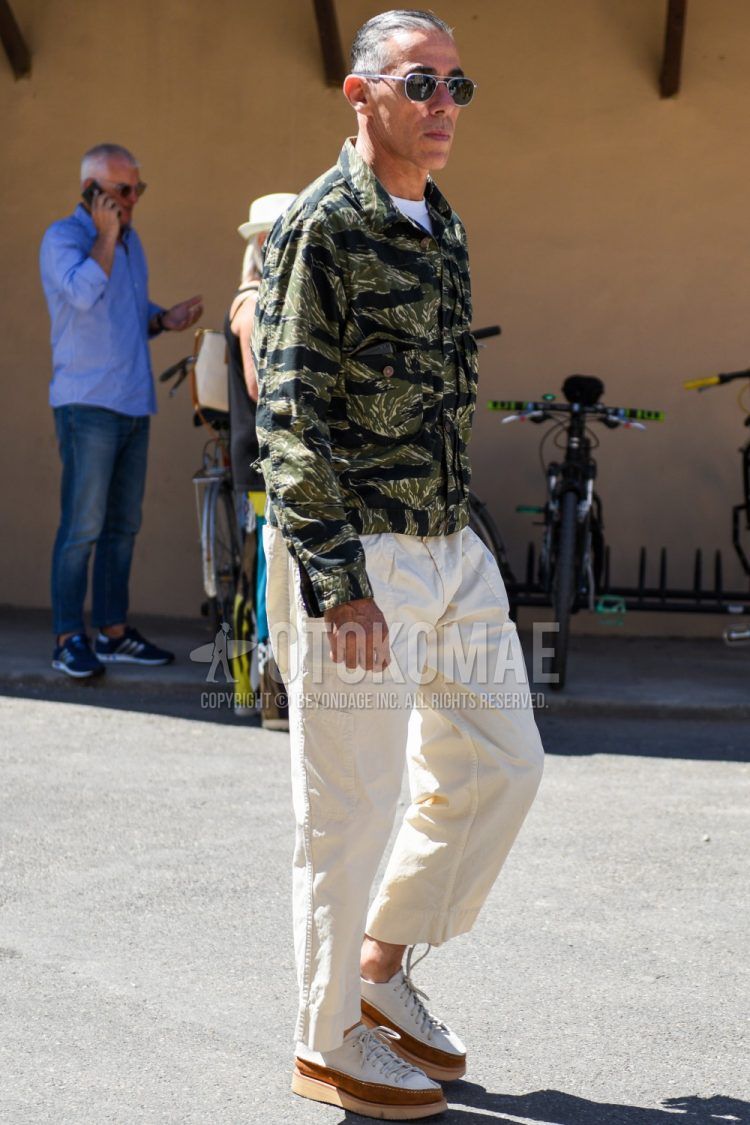 Men's coordinate and outfit with plain black and silver sunglasses, olive green and black camouflage shirt jacket, plain beige chinos, plain pleated pants, and white and brown leather shoes.