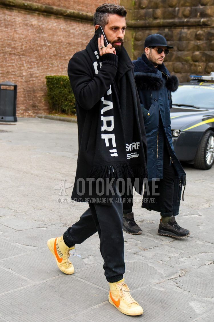 Men's coordinate and outfit with black graphic scarf/stall, solid black outerwear, solid black jogger pants/ribbed pants, and Nike orange high-cut sneakers.