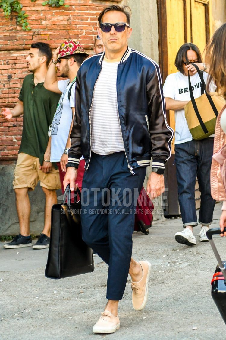 Men's outfit/combination of plain sunglasses, plain black stadium jacket, white/gray striped t-shirt, plain navy cropped pants, plain pleated pants, Vans beige low-cut sneakers, and plain black briefcase/handbag.