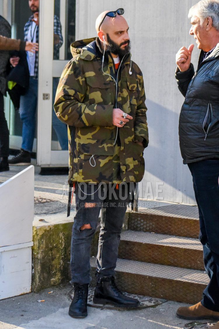 Men's coordinate and outfit with plain black sunglasses, olive green/brown camouflage down jacket, plain white t-shirt, plain gray damaged jeans, and black boots.