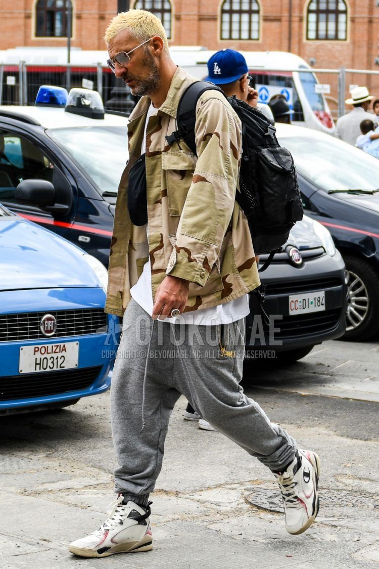 Men's coordinate and outfit with plain gold sunglasses, beige camouflage field jacket/hunting jacket, plain white t-shirt, plain gray sweatpants, Champion white high-cut sneakers, plain black backpack, and plain black body bag.