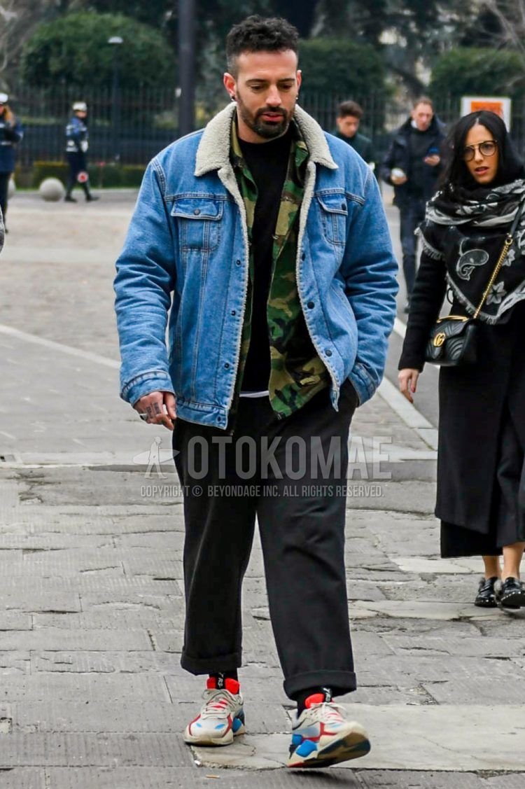 Men's coordinate and outfit with solid blue denim jacket, green camouflage shirt jacket, solid black sweater, dark gray solid slacks, and multi-colored low-cut sneakers.