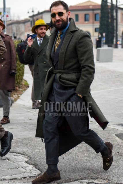 Men's coordinate and outfit with solid color sunglasses, olive green solid color belted coat, dark gray solid color socks, brown monk shoes leather shoes, brown suede shoes leather shoes, dark gray striped suit, yellow tie tie.