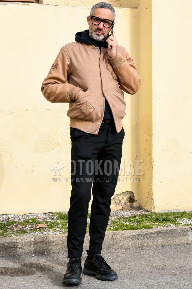 Men's coordinate and outfit with solid color glasses, solid color brown stadium jacket, solid color black hoodie, solid color black slacks, solid color black socks, and black low-cut sneakers.