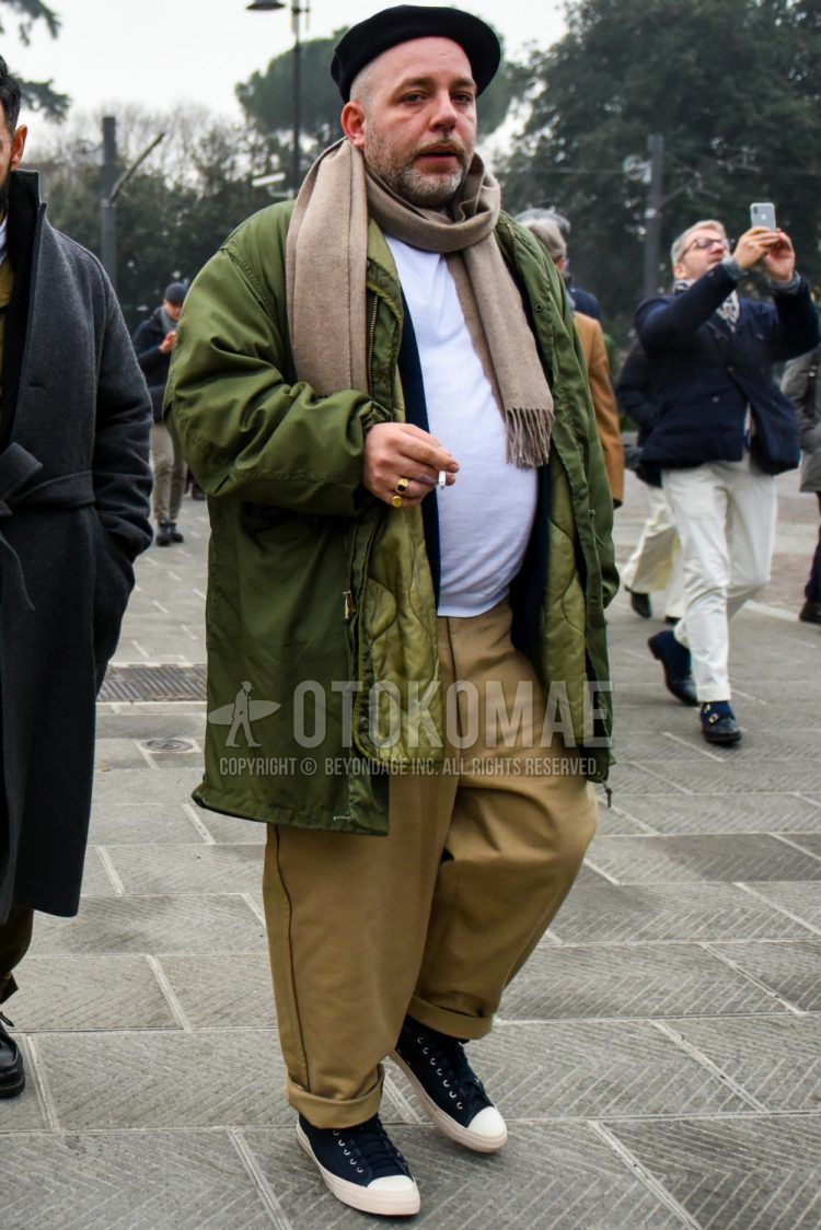 Men's outfit with solid black cap, solid brown scarf/stall, solid olive green outerwear, solid olive green quilted jacket, solid white t-shirt, solid beige wide-leg pants, solid beige chinos, navy low-cut sneakers. Outfit.