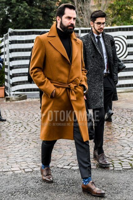 Men's coordinate and outfit with solid beige belted coat, solid black turtleneck knit, solid gray slacks, blue dot socks, and brown straight tip leather shoes.