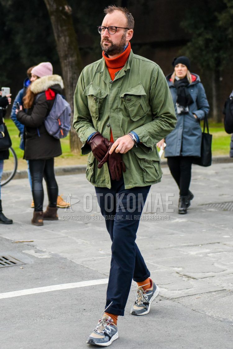 Men's coordinate and outfit with solid color glasses, solid color green safari jacket, solid color orange turtleneck knit, solid color blue denim/chambray shirt, solid color gray cotton pants, solid color orange socks, and gray low cut sneakers.