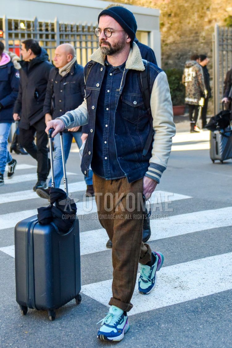 Men's coordinate and outfit with solid black knit cap, solid black glasses, solid blue denim jacket, solid blue denim/chambray shirt, brown checked slacks, Adidas Falcon multi-colored low-cut sneakers, and solid dark gray suitcase. .