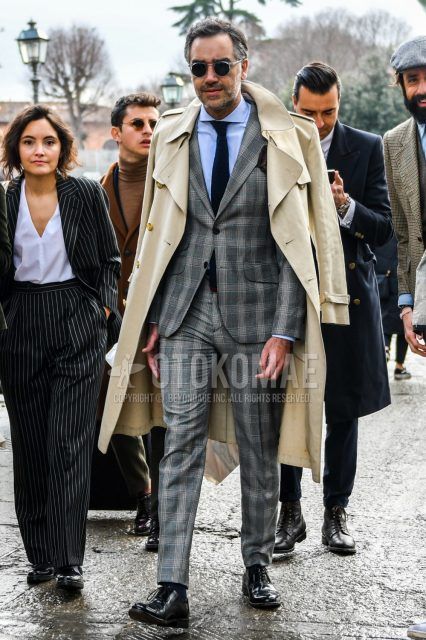 Men's winter/autumn coordinate and outfit with clear solid color sunglasses, solid color beige trench coat, solid color light blue shirt, black plain toe leather shoes, gray checked suit, and solid color navy knit tie.