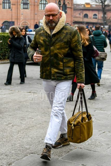 Men's coordinate and outfit with brown tortoiseshell glasses, olive green camouflage leather jacket (not riders), plain white cotton pants, white/black striped socks, brown low-cut New Balance sneakers, and plain beige briefcase/handbag.