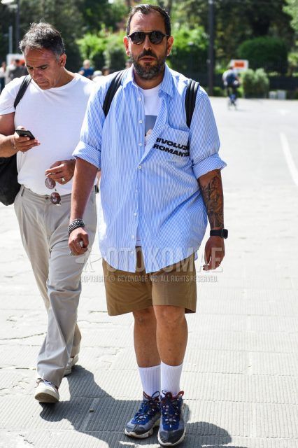 Men's coordinate and outfit with plain black sunglasses, light blue/white striped shirt, plain white t-shirt, plain beige shorts, plain white socks, and gray/blue low-cut sneakers.