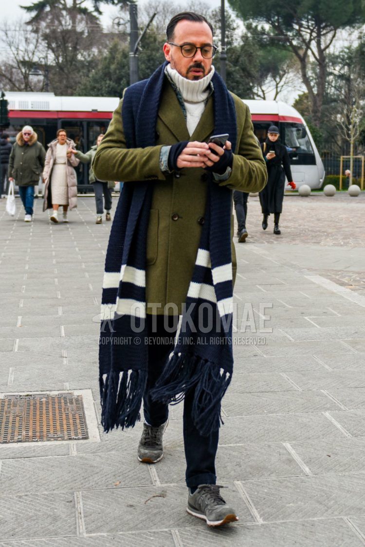 Men's coordinate and outfit with plain black glasses, plain navy scarf/stall, plain olive green chester coat, plain white turtleneck knit, plain navy chinos, and New Balance gray low-cut sneakers.