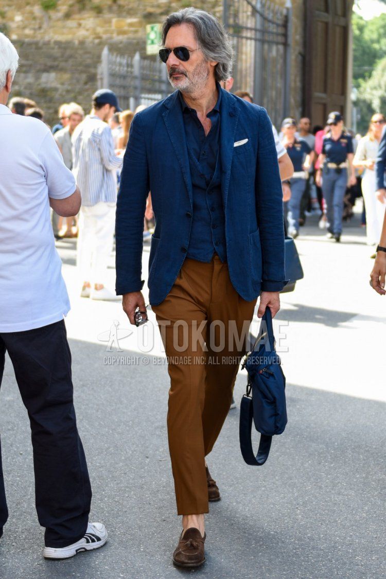Men's outfit/clothing with plain black sunglasses, plain navy tailored jacket, plain navy shirt, plain navy gilet, plain brown slacks, suede brown tassel loafer leather shoes, and plain blue briefcase/handbag.