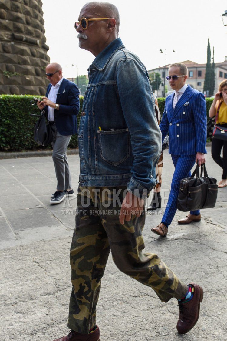 Men's coordinate and outfit with solid yellow sunglasses, solid blue denim jacket, green camouflage cotton pants, green camouflage wide-leg pants, multi-colored solid socks, and brown brogue shoes leather shoes.