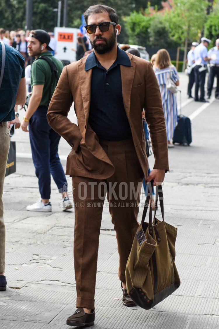 Men's spring/summer/fall outfit with plain black Wellington sunglasses, plain navy polo shirt, suede brown loafer leather shoes, plain beige tote bag, and plain brown suit.