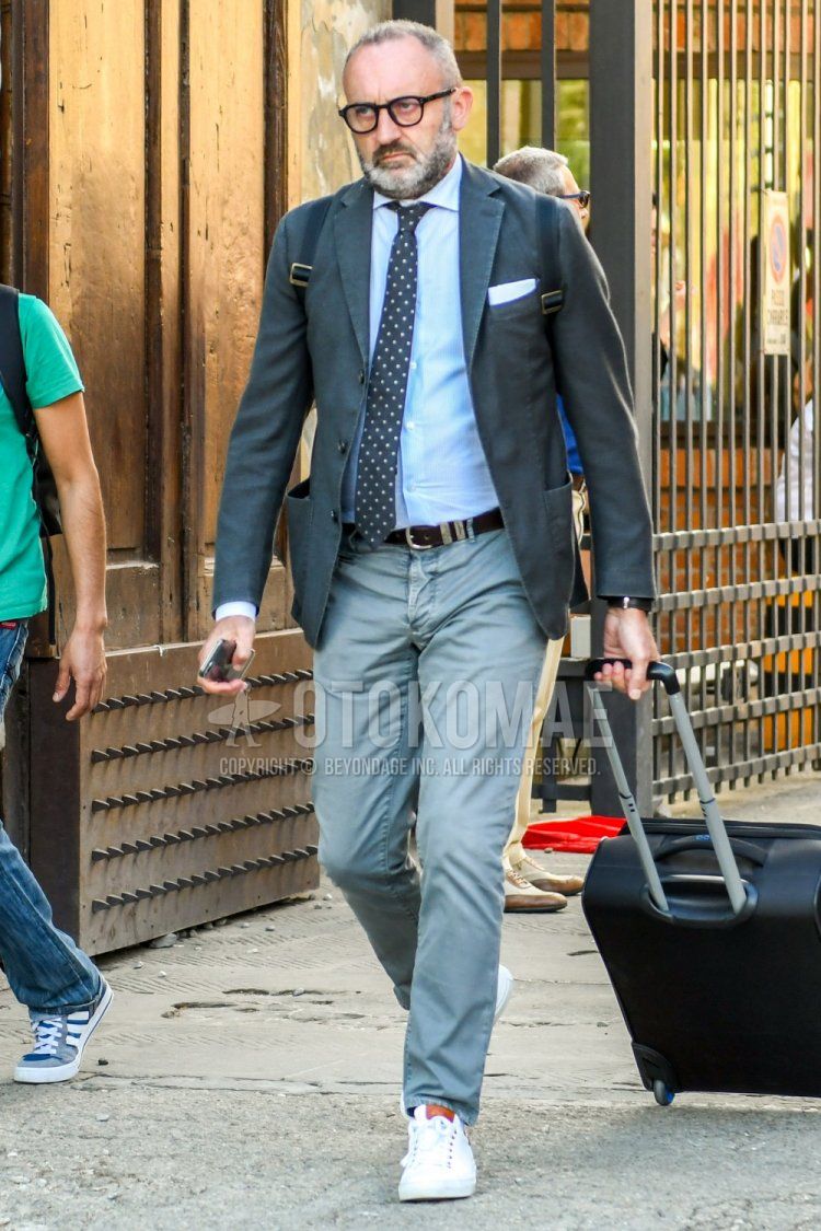 Men's coordinate and outfit with plain glasses, dark gray plain tailored jacket, light blue plain shirt, brown plain leather belt, gray plain chinos, white low-cut sneakers, plain bag, and navy dot tie.