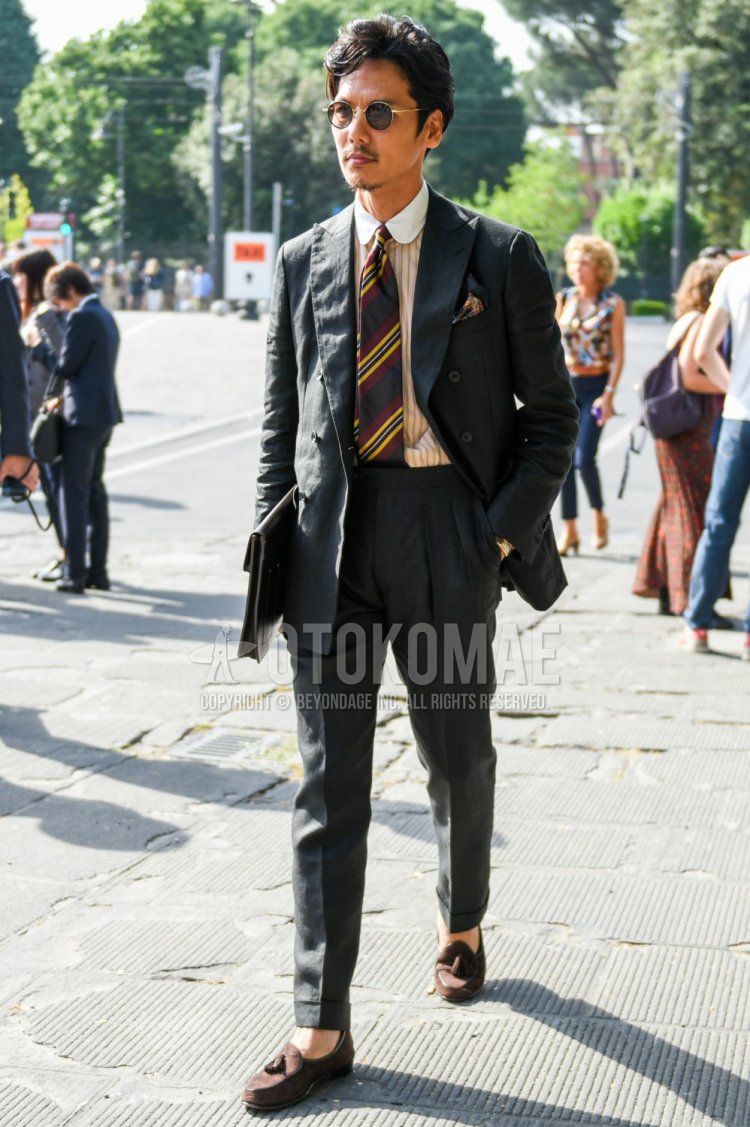 Men's coordinate and outfit with solid gold sunglasses, yellow striped shirt, brown tassel loafers leather shoes, brown suede shoes leather shoes, solid black clutch bag/second bag/drawstring, solid dark gray suit, and multicolor regimental tie.