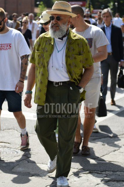 Men's coordinate and outfit with solid beige hat, solid silver sunglasses, olive green botanical shirt, solid white t-shirt, solid black leather belt, solid green wide-leg pants, and white low-cut sneakers.