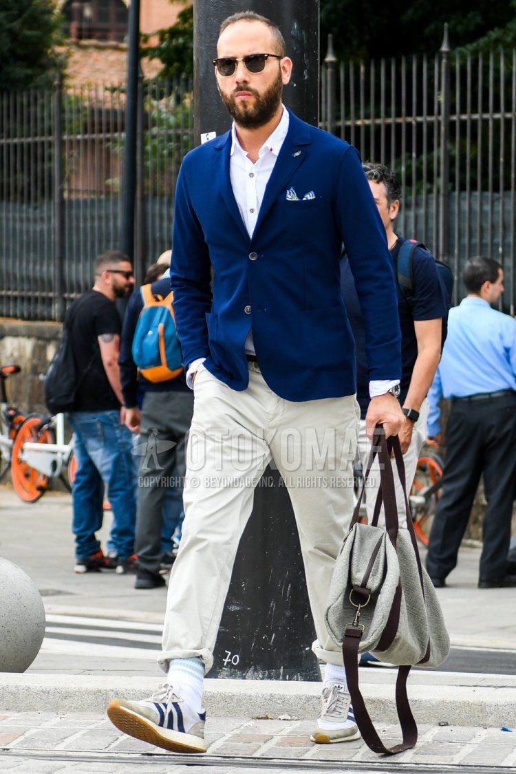 Men's outfit with plain sunglasses, plain navy tailored jacket, plain white shirt, plain white cotton pants, white striped socks, Adidas beige low-cut sneakers, and plain gray briefcase/handbag.