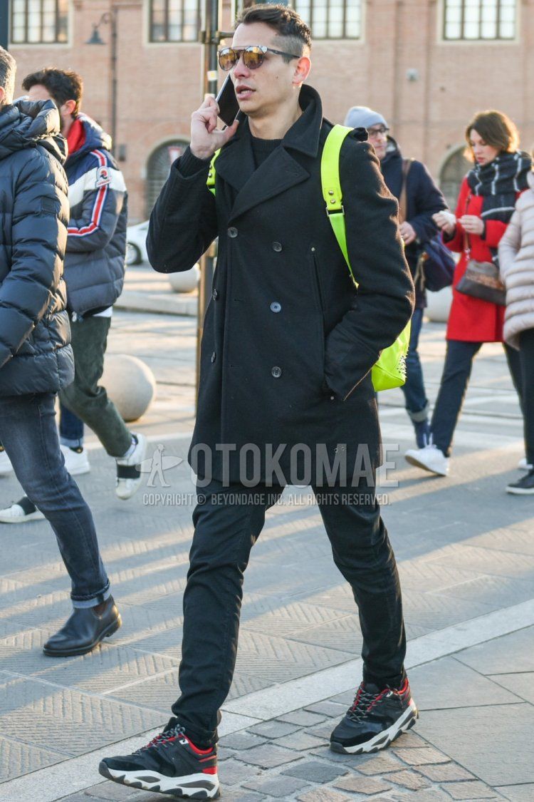 Men's fall/winter outfit with plain silver sunglasses, plain dark gray P-coat, plain black sweater, plain dark gray denim/jeans, black low-cut sneakers, and plain yellow backpack.