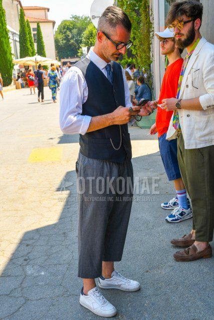 Men's coordinate and outfit with plain glasses, dark gray solid color gilet, white solid color shirt, gray solid color wide pants, gray solid color cropped pants, Diadora white low-cut sneakers, and blue solid color tie.