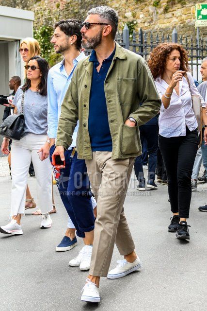 Men's coordinate and outfit with solid color sunglasses, olive green solid color shirt jacket, navy solid color polo shirt, beige solid color ankle pants, and white low-cut sneakers.