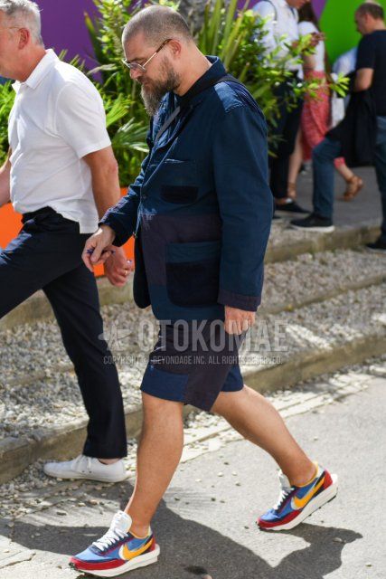 Men's coordinate and outfit with plain silver sunglasses, plain navy tailored jacket, plain navy t-shirt, plain navy shorts, and Nike Sakai LD Waffle red, light blue, and yellow low-cut sneakers.