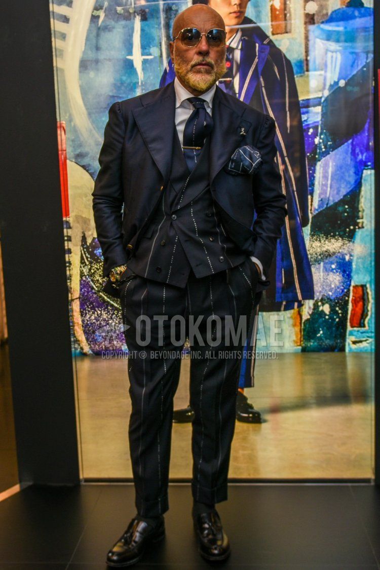 Men's spring and fall outfit with plain black and gold sunglasses, plain navy tailored jacket, navy striped gilet, navy striped slacks, plain gray socks, black tassel loafer leather shoes, and navy striped tie.