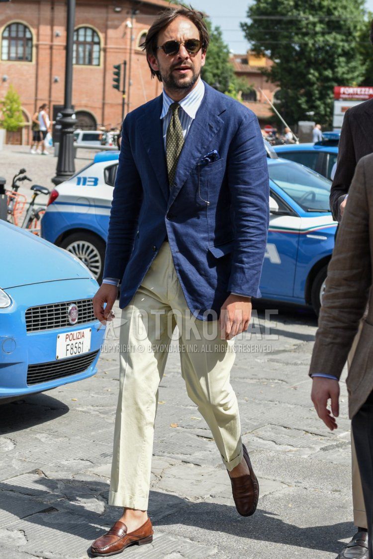 Men's coordinate and outfit with plain black sunglasses, plain navy tailored jacket, white/light blue striped shirt, plain beige cotton pants, brown coin loafer leather shoes, olive green dot tie.