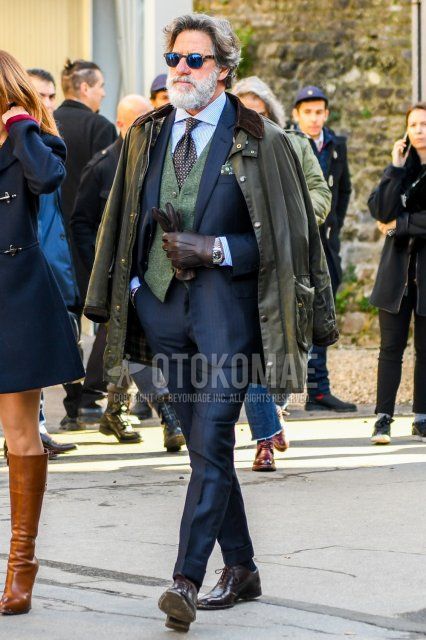 Men's coordinate and outfit with plain sunglasses, plain olive green field jacket/hunting jacket, plain green gilet, white and light blue striped shirt, plain black socks, brown straight tip leather shoes, plain gray suit, and brown small print tie.