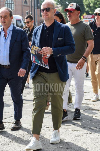 Men's spring/summer/fall outfit with plain gray sunglasses, plain navy tailored jacket, plain navy polo shirt, solid olive green cotton pants, solid olive green cropped pants, and white low-cut sneakers.
