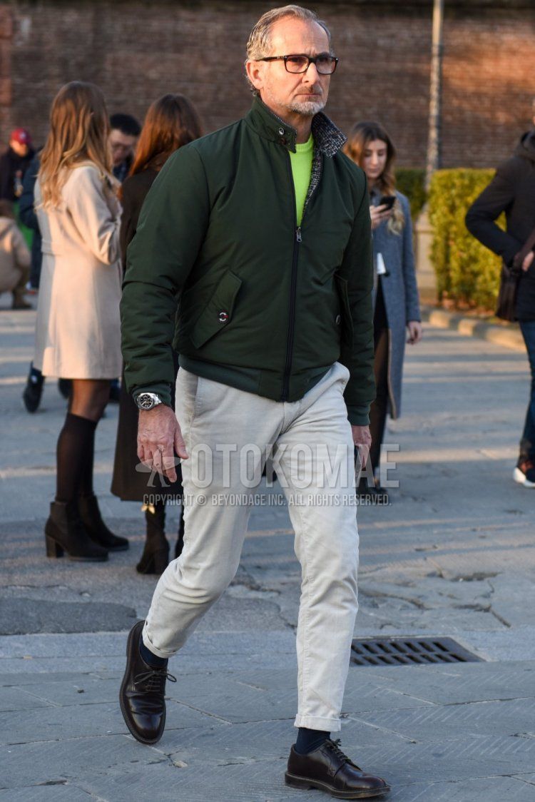 Men's fall/winter outfit and outfit with solid black glasses, solid olive green outerwear, solid yellow sweater, solid beige chinos, solid navy socks, and black plain toe leather shoes.