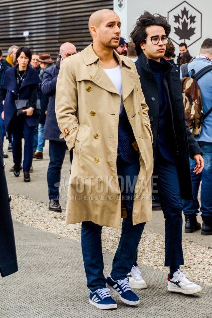 Men's coordinate and outfit with plain beige trench coat, plain white t-shirt, plain navy tailored jacket, plain navy denim/jeans, and navy low-cut Adidas sneakers.
