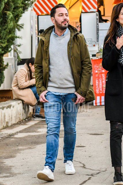 Men's coordinate and outfit with olive green solid color mountain parka, gray solid color sweater, light blue solid color shirt, blue solid color denim/jeans, and white low-cut sneakers.