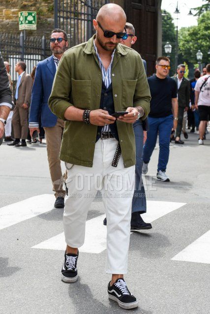 Men's coordinate and outfit with Wellington plain black sunglasses, olive green plain shirt jacket, light blue and white striped shirt, navy plain gilet, black dotted leather belt, plain white ankle pants, and Vans black low-cut sneakers.