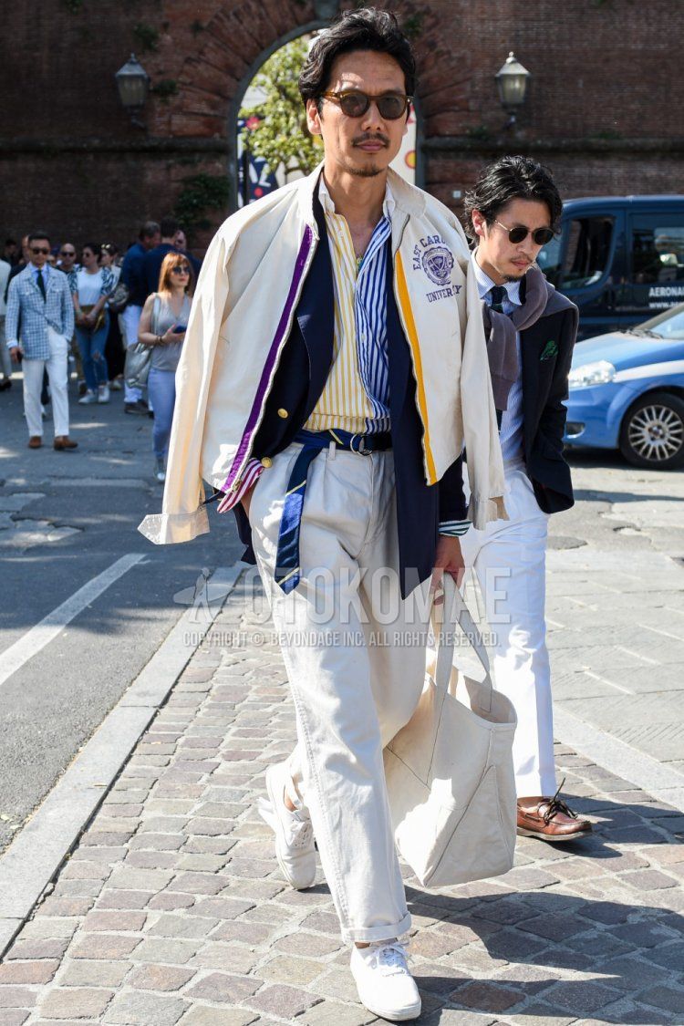 Men's outfit/clothing with plain brown sunglasses, plain beige outerwear, plain navy tailored jacket, yellow/navy striped shirt, plain navy tape belt, plain white cotton pants, white low-cut sneakers, and plain white tote bag.