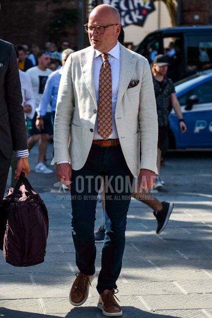Men's coordination and outfit with black tortoiseshell glasses, plain beige/white tailored jacket, plain white shirt, plain brown leather belt, plain navy cotton pants, brown low-cut sneakers, and red tie tie.