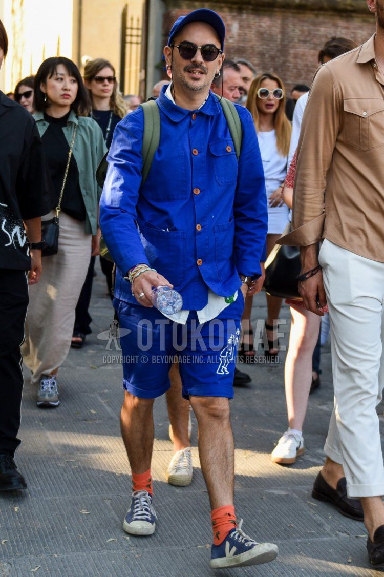 Men's coordinate and outfit with plain blue baseball cap, plain black Boston sunglasses, plain blue shirt jacket, plain white shirt, blue graphic shorts, plain orange socks, and navy low-cut sneakers.