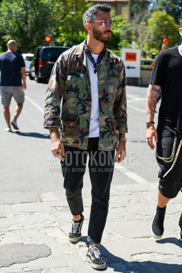 Men's coordinate and outfit with teardrop gold plain glasses, black paisley bandana/neckerchief, green camouflage safari jacket, plain white t-shirt, plain black denim/jeans, and black low-cut sneakers by Vans.