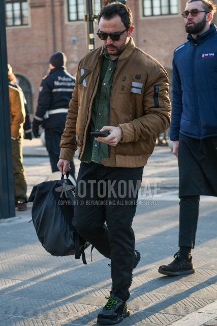 Men's fall/winter outfit with solid black sunglasses, solid beige MA-1, solid olive green shirt, dark gray solid slacks, black low-cut sneakers, and solid gray backpack.