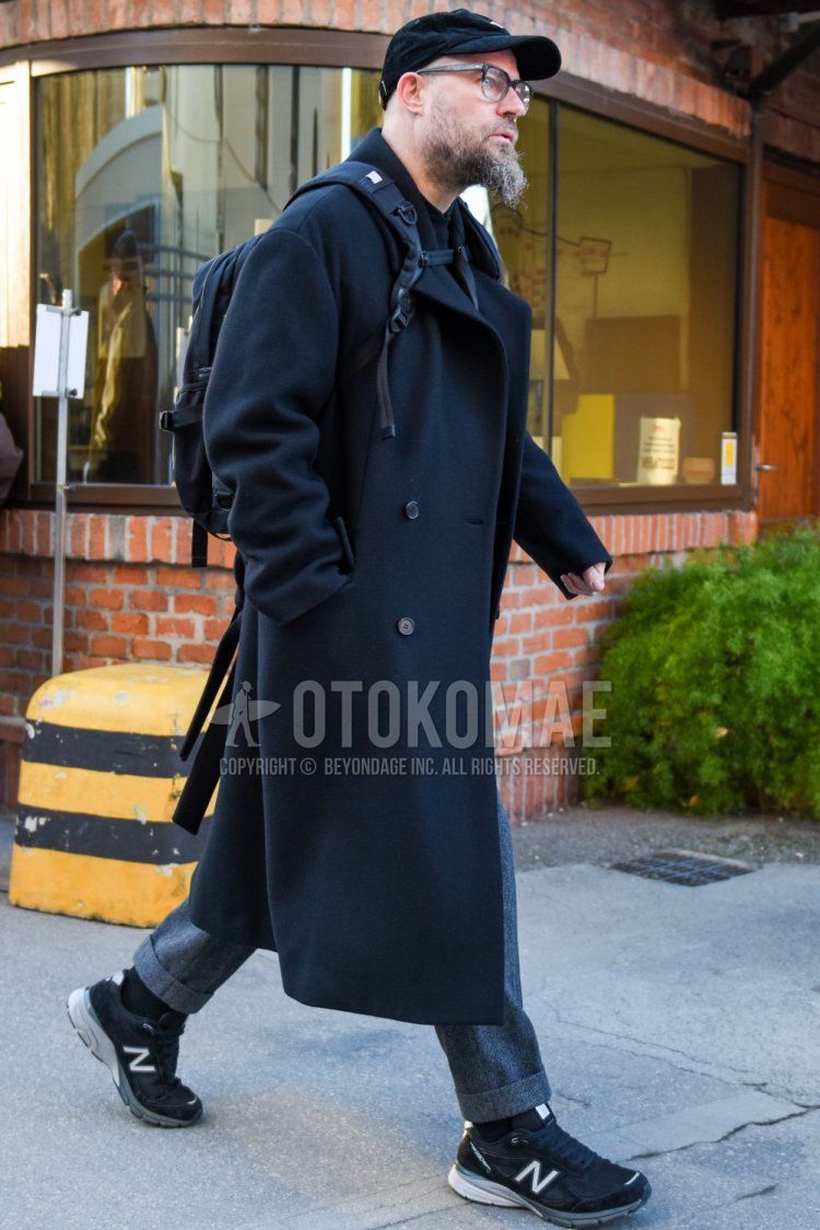 Men's fall/winter outfit and outfit with black one-point baseball cap, solid black glasses, solid black trench coat, solid gray slacks, solid black socks, New Balance 990 black low-cut sneakers, and solid black backpack.