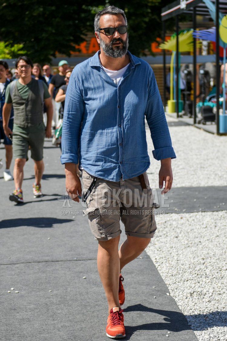 Men's spring/summer coordinate and outfit with Wellington solid black sunglasses, solid light blue denim/chambray shirt, white graphic t-shirt, solid beige cargo pants, solid shorts, and Nike red low-cut sneakers.