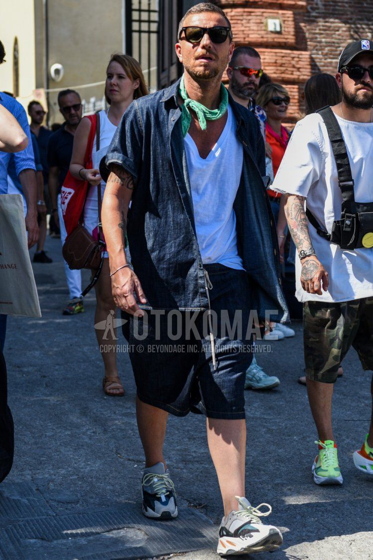 Summer men's coordinate and outfit with solid black sunglasses, solid green bandana/neckerchief, solid white t-shirt, solid navy denim/chambray shirt, solid navy shorts, and multi-colored low-cut sneakers.
