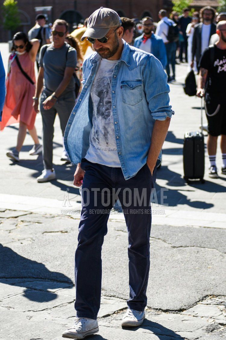 Men's spring/summer coordinate and outfit with plain gray baseball cap, plain black/silver sunglasses, plain blue denim/chambray shirt, white graphic t-shirt, plain navy chinos, and Converse All Star white high-cut sneakers.