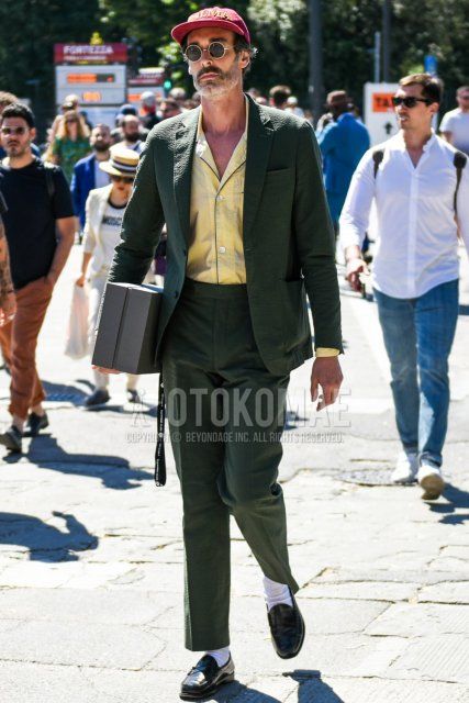 Men's spring and summer coordinate and outfit with red decal logo baseball cap, round black and gold solid sunglasses, open collar solid yellow shirt, solid white socks, black coin loafer leather shoes, and olive green solid suit.