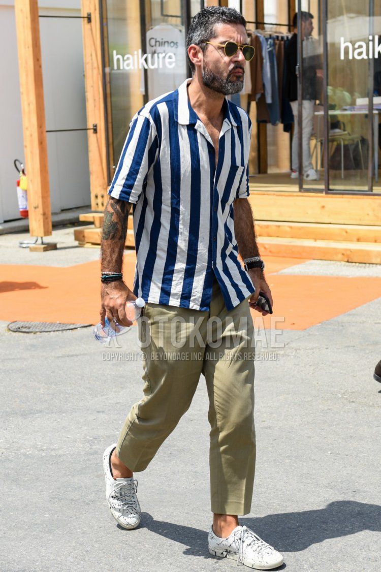Summer men's coordinate and outfit with gold and black solid color sunglasses, white and navy striped shirt, beige solid color ankle pants and white low-cut sneakers.