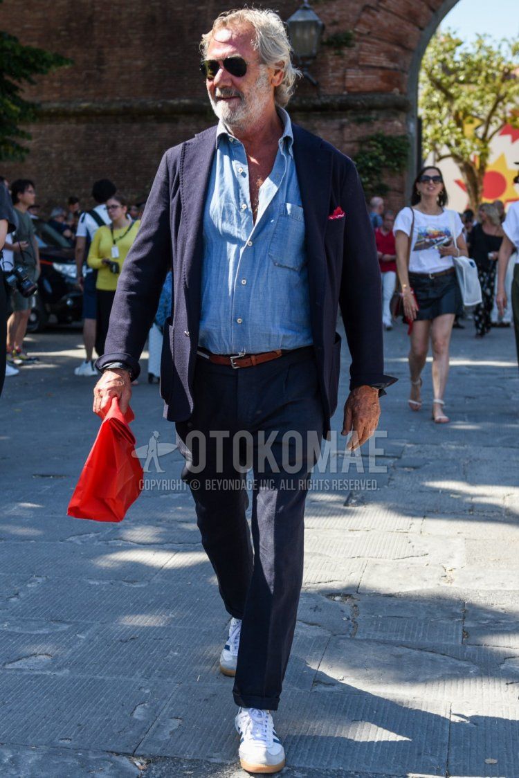 Men's spring/summer/fall outfit with gold/black solid color sunglasses, navy solid color tailored jacket, light blue solid color denim/chambray shirt, brown solid color leather belt, navy solid color slacks, and white low-cut Adidas sneakers.
